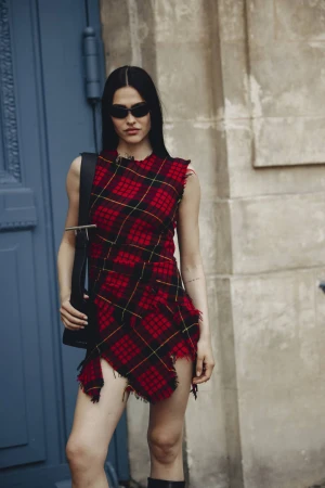 Model in red plaid dress and dark sunglasses holding a black sling bag standing against a stone building exterior