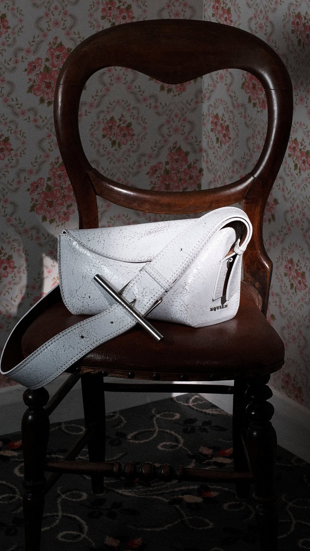 White leather sling bag rests on a dark wooden vintage chair against a floral patterned wallpaper background