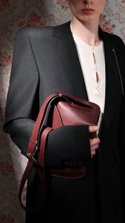 Model holding a structured burgundy leather sling bag wearing a black coat and white top against a floral wallpaper