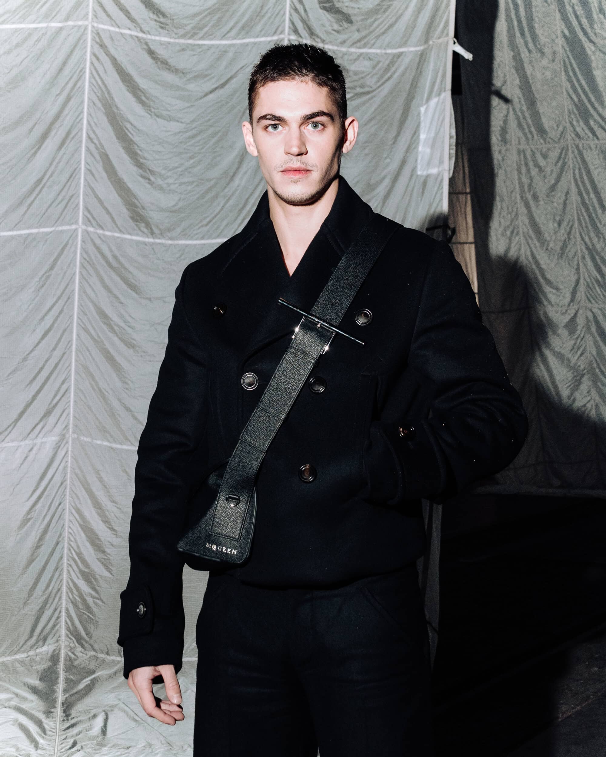 A man wearing a black double-breasted pea coat and a black sling bag standing in front of a wrinkled fabric backdrop.
