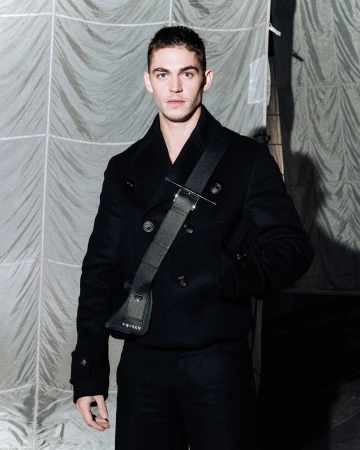 A man wearing a black double-breasted pea coat and a black sling bag standing in front of a wrinkled fabric backdrop.
