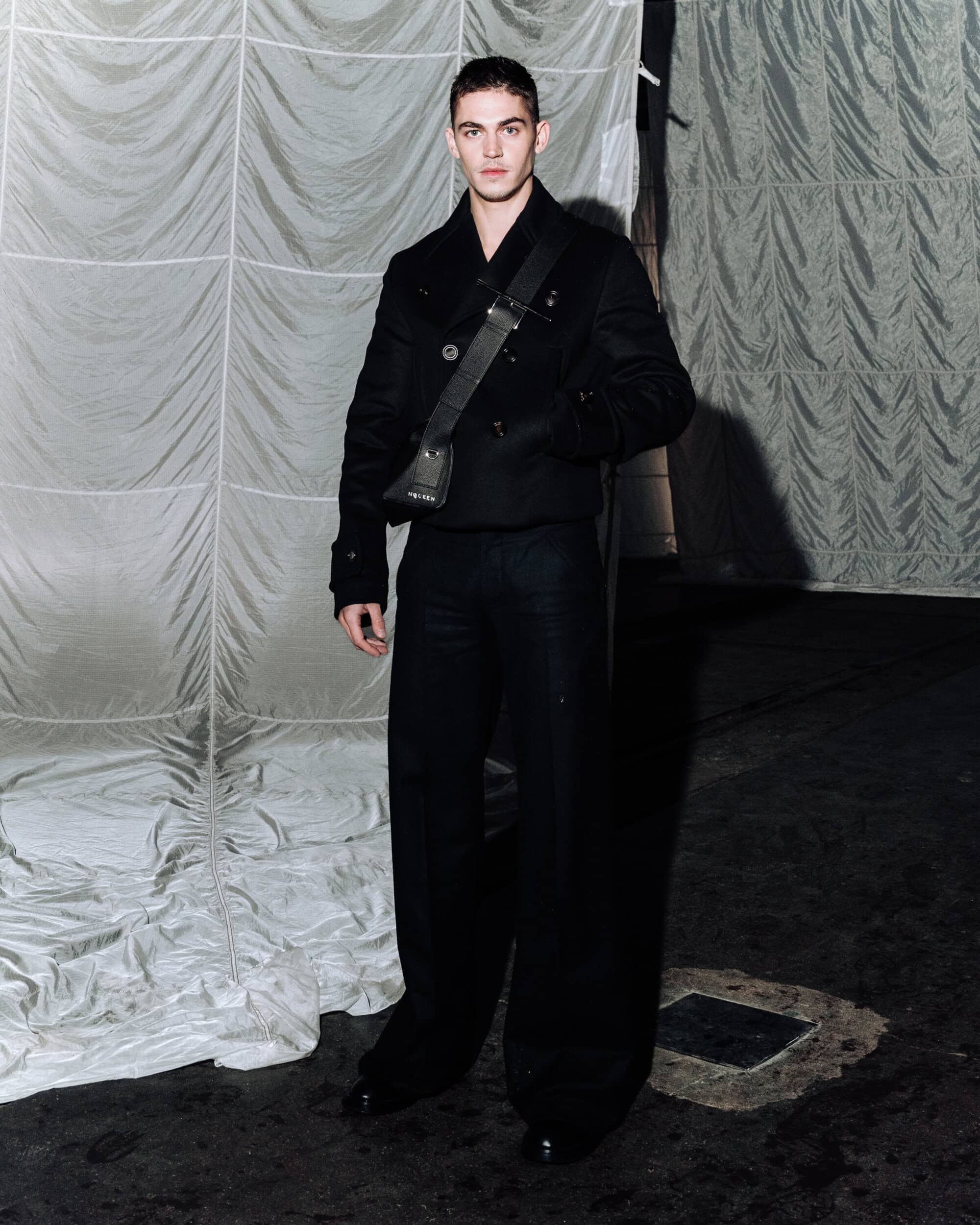 Man wearing a black jacket and carrying a sling bag against a draped backdrop for McQueen