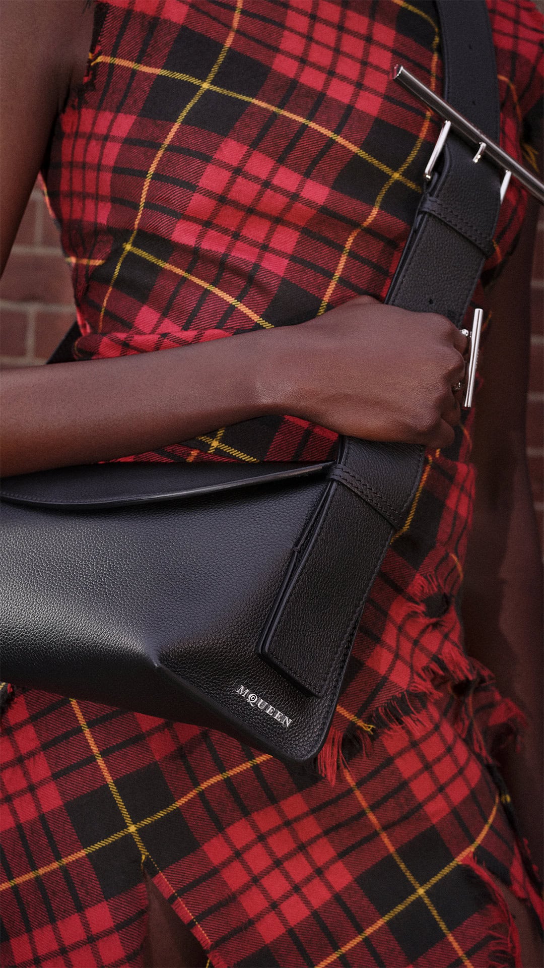 Close-up of a person wearing a red plaid dress holding a black leather sling bag from McQueen.