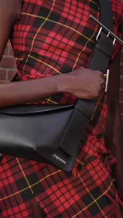 Close-up of a person wearing a red plaid dress holding a black leather sling bag from McQueen.