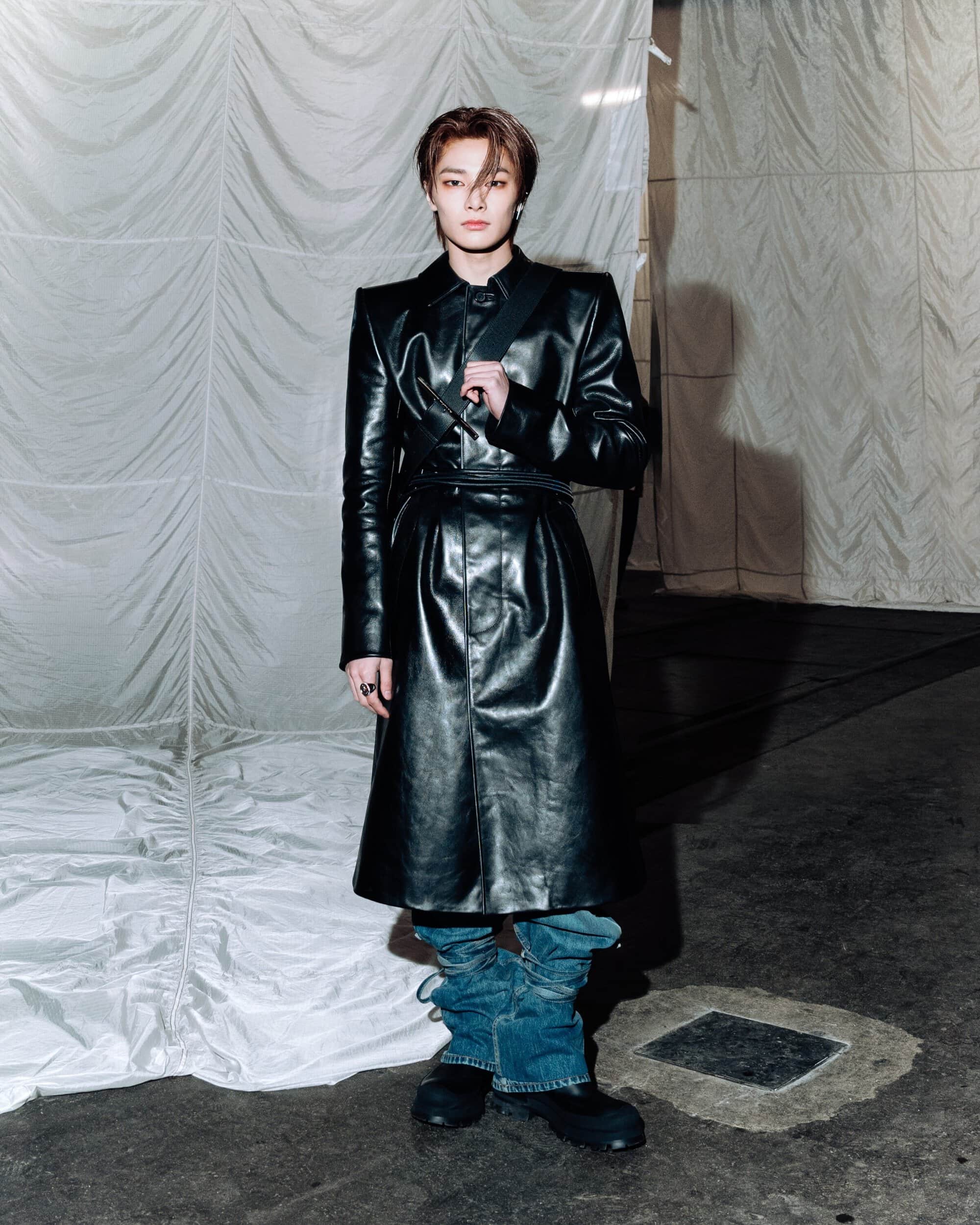A man wearing a black leather coat and denim boots standing against a draped white fabric backdrop.