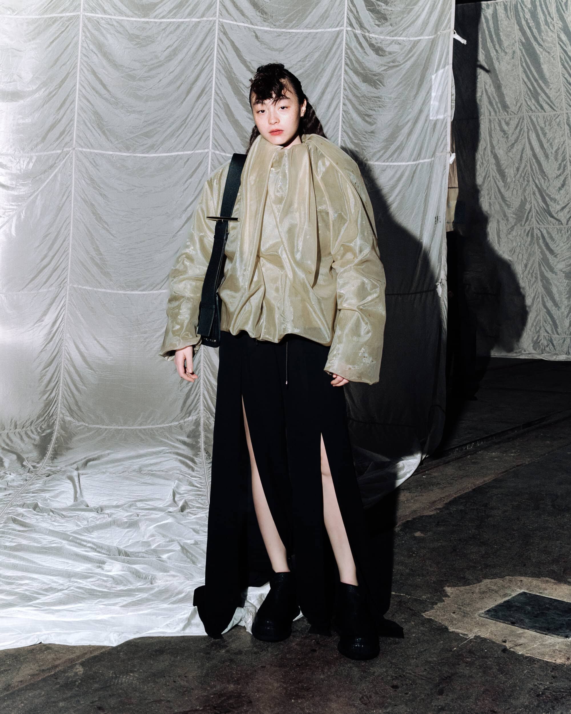 Model wearing an olive green jacket and black slit skirt carrying a black sling bag against a fabric backdrop.