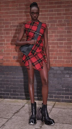 Model standing against a red brick wall wearing a red plaid dress and black sling bag for McQueen campaign