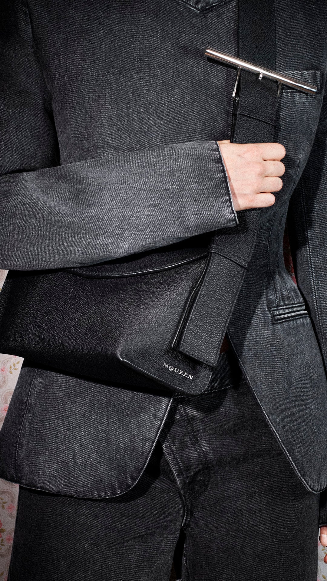 Close-up of a person wearing a dark denim jacket holding a black leather sling bag with silver hardware.