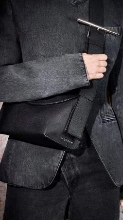 Close-up of a person wearing a dark denim jacket holding a black leather sling bag with silver hardware.