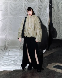 Model wearing an olive green jacket and black slit skirt carrying a black sling bag against a fabric backdrop.
