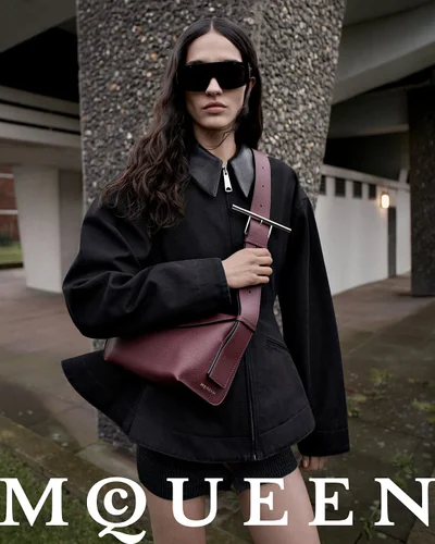 Model wearing dark sunglasses and a black jacket carrying a burgundy crossbody sling bag outdoors.
