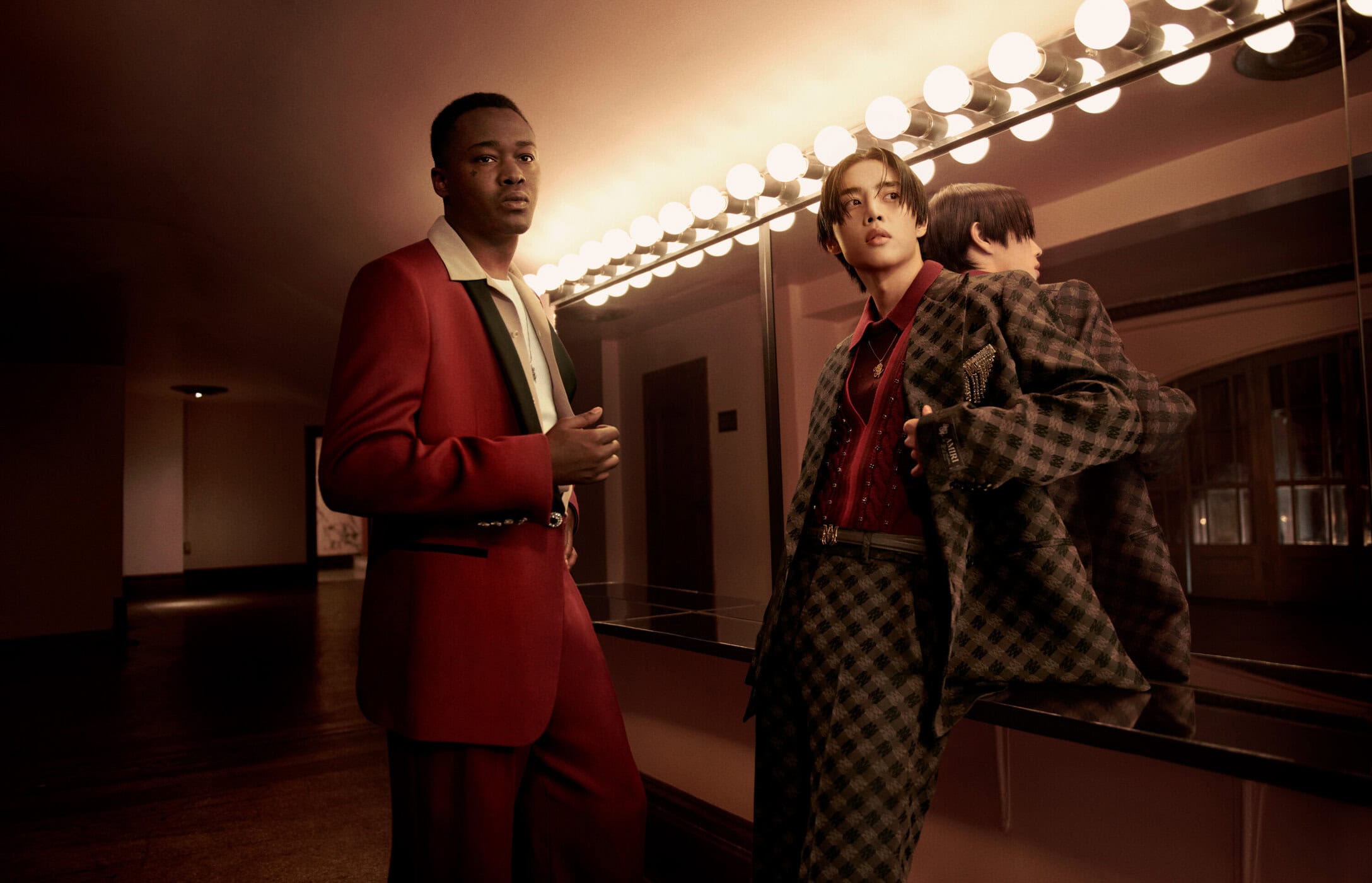 Two men in tailored suits posing in front of a mirror with warm vanity lighting for Amiri Fall 2024 ad campaign.