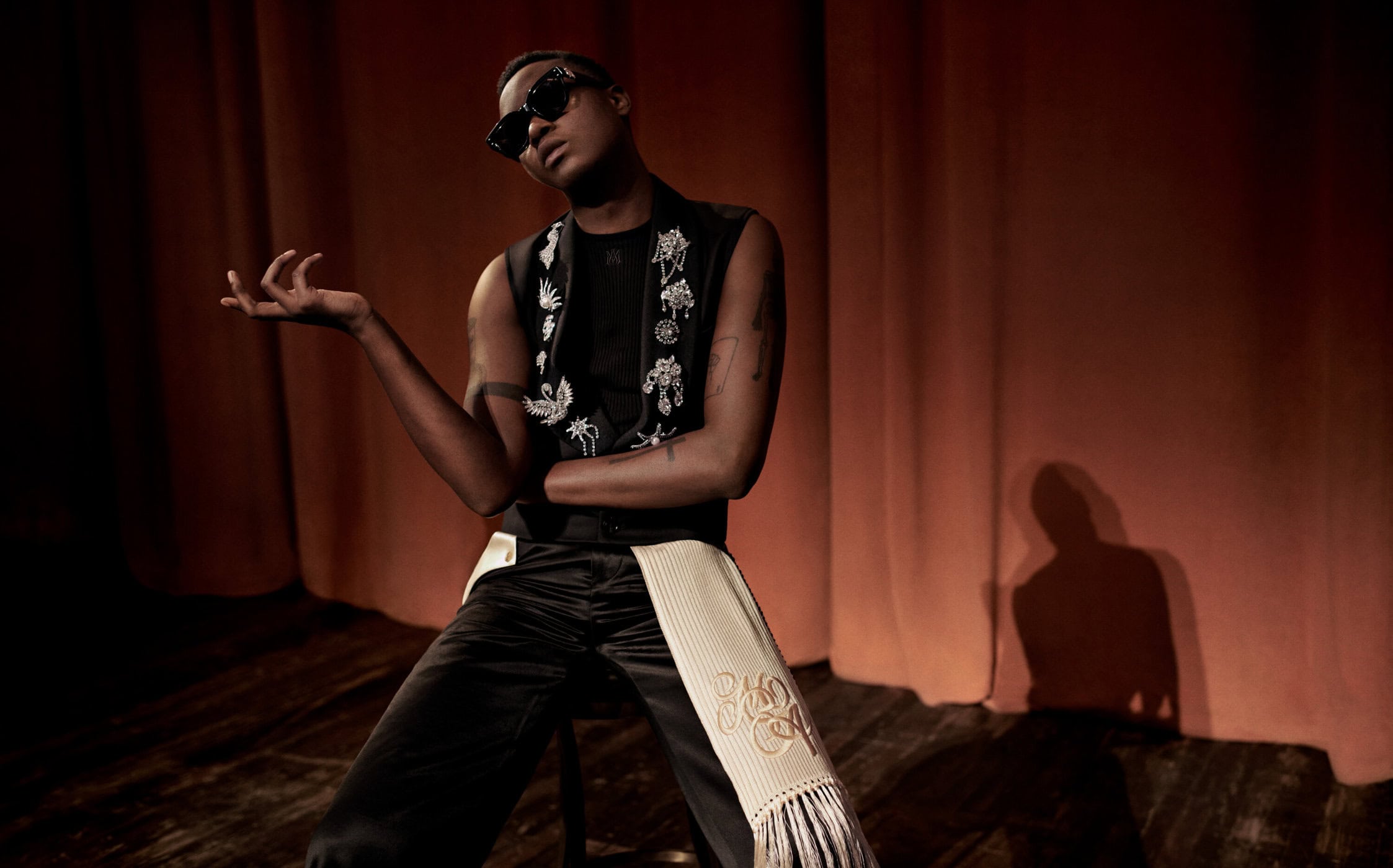 Model wearing dark sunglasses and embellished vest posing in front of gathered curtains for Amiri campaign