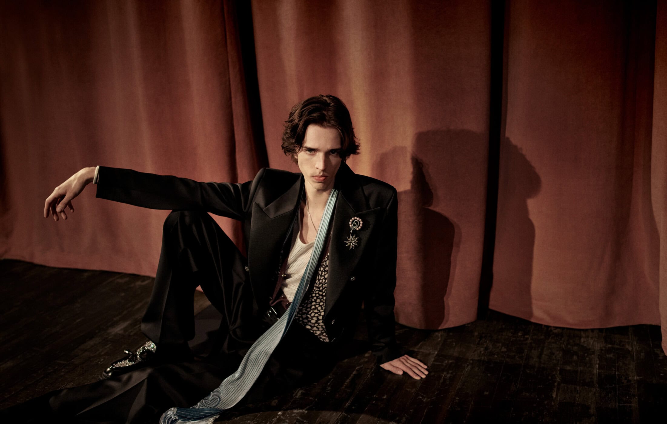 A young man in a black suit and tie sitting against heavy red curtains for the Amiri Fall 2024 campaign