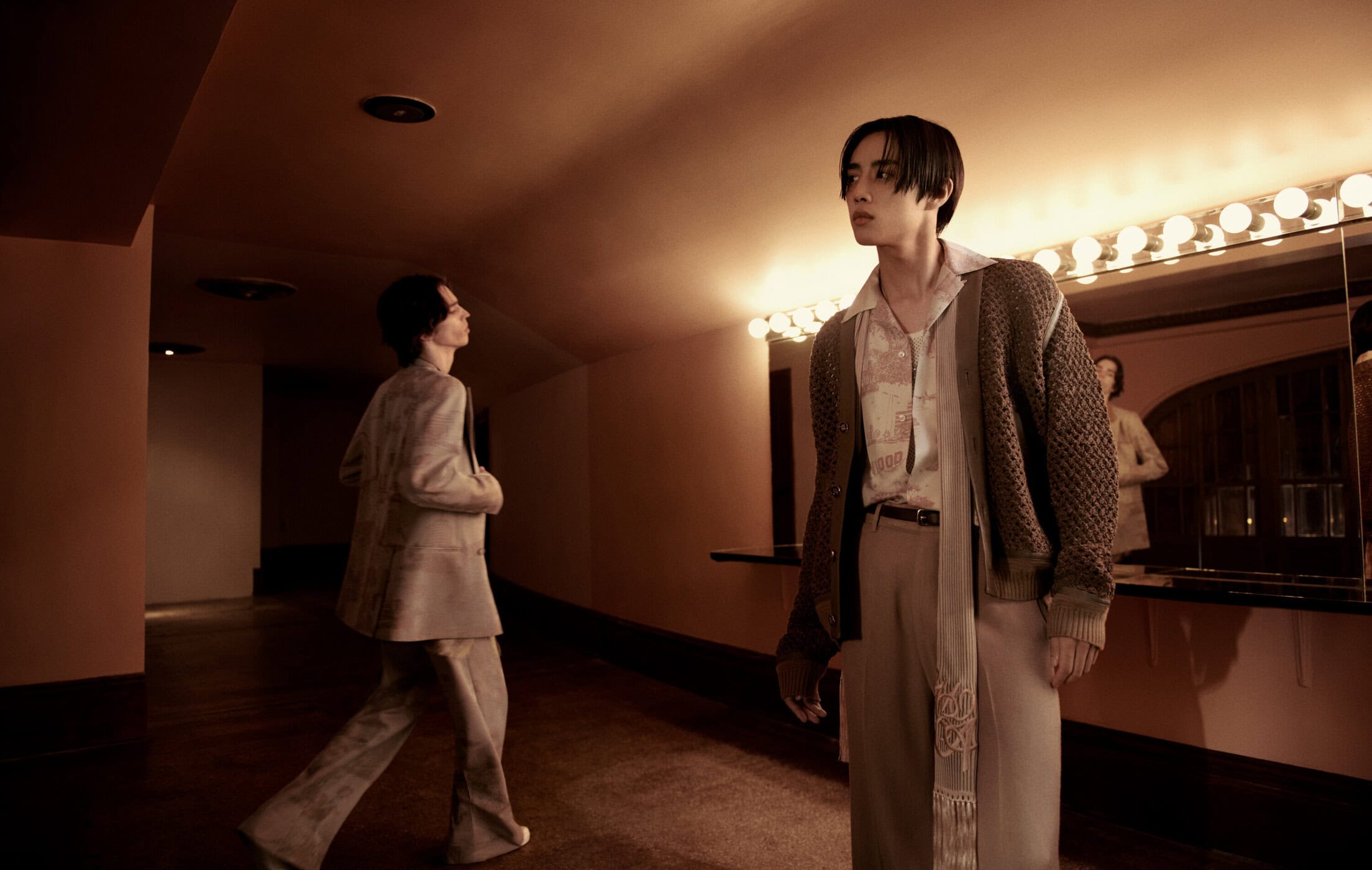 Two men modeling Amiri Fall 2024 collection in a warm-toned interior with dressing room mirror lights