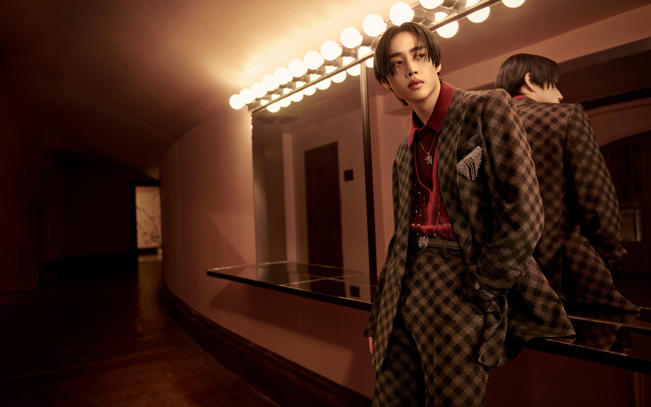 Man wearing a brown plaid suit and red shirt leaning against a mirror with Hollywood vanity lights in a hallway.