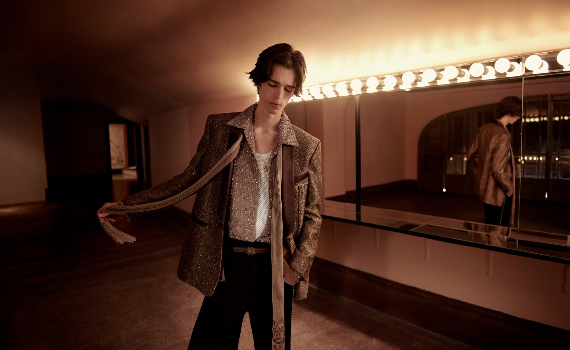Model in a brown blazer holding a long scarf standing in front of a lit vanity mirror