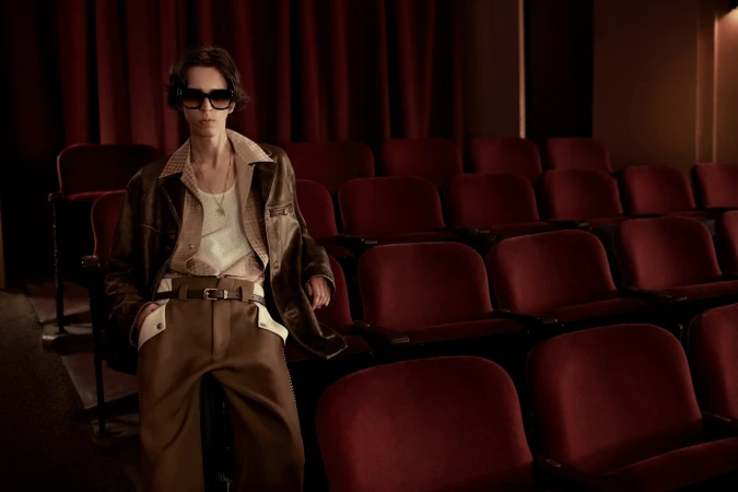 Model wearing dark sunglasses, brown leather jacket, and trousers seated in a vintage cinema with red velvet seats.