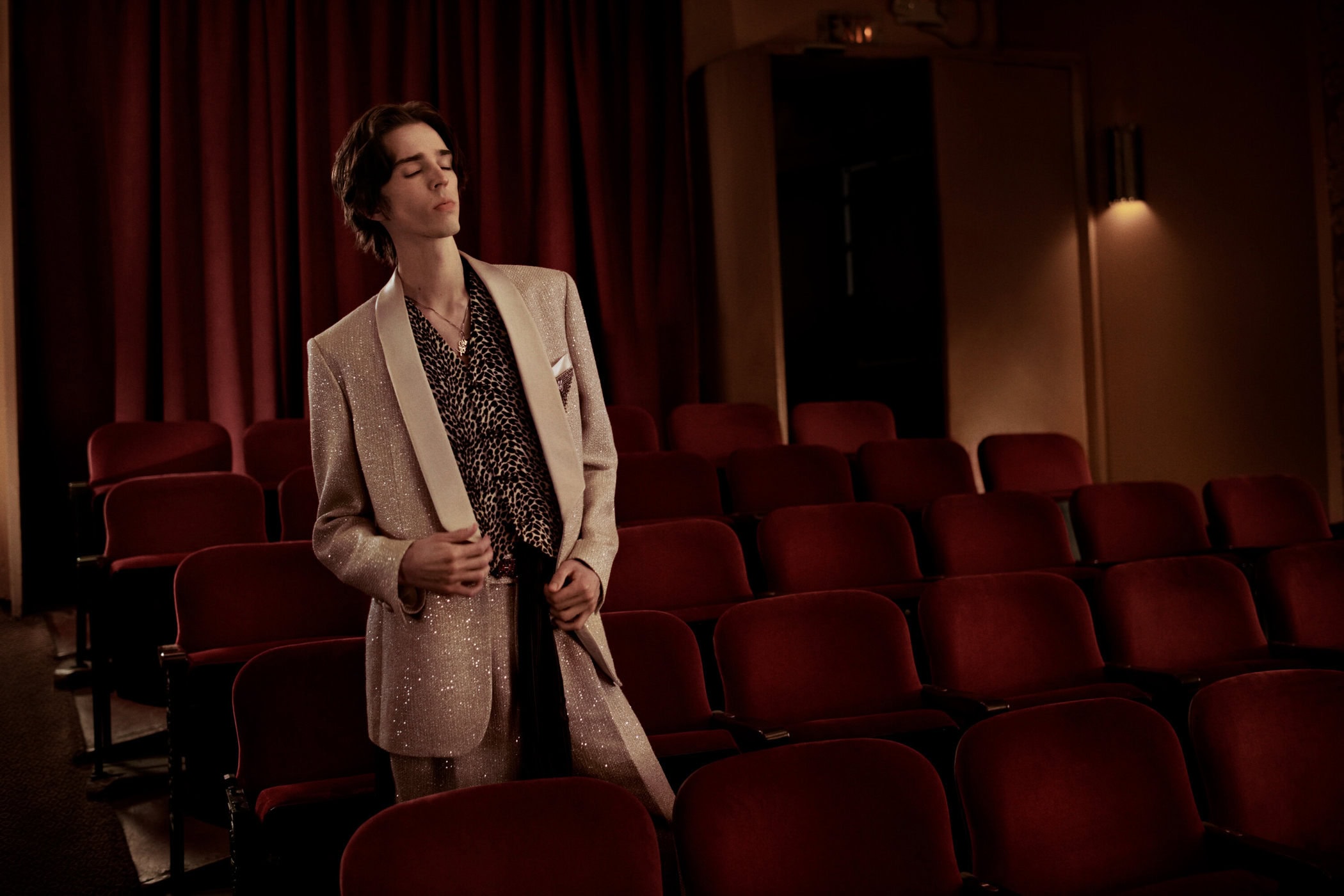 A man in a shimmering cream blazer and animal print shirt stands in an empty movie theater with red velvet seats.