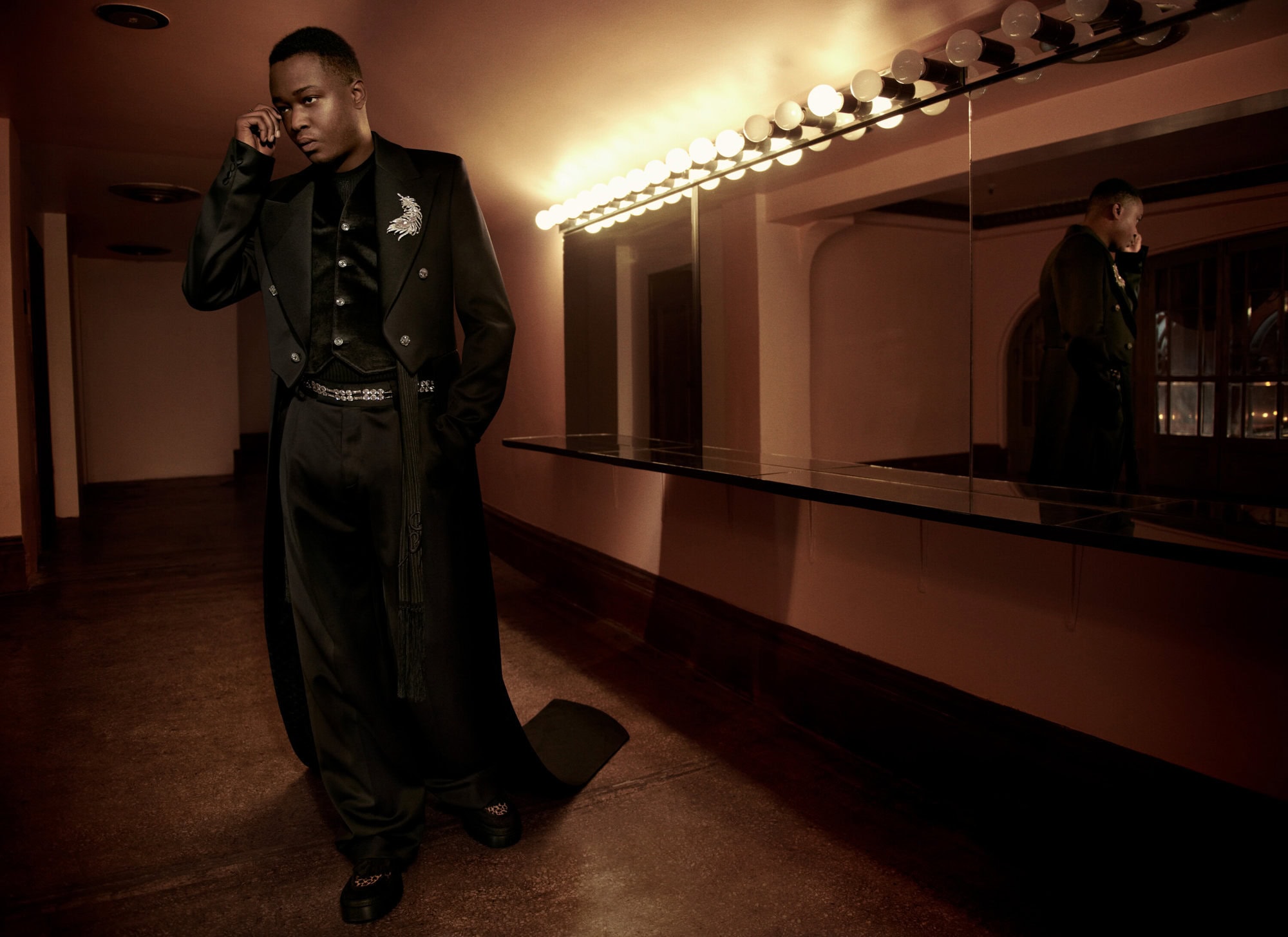 Man wearing a tailored black coat and matching trousers standing in a warmly lit backstage dressing room