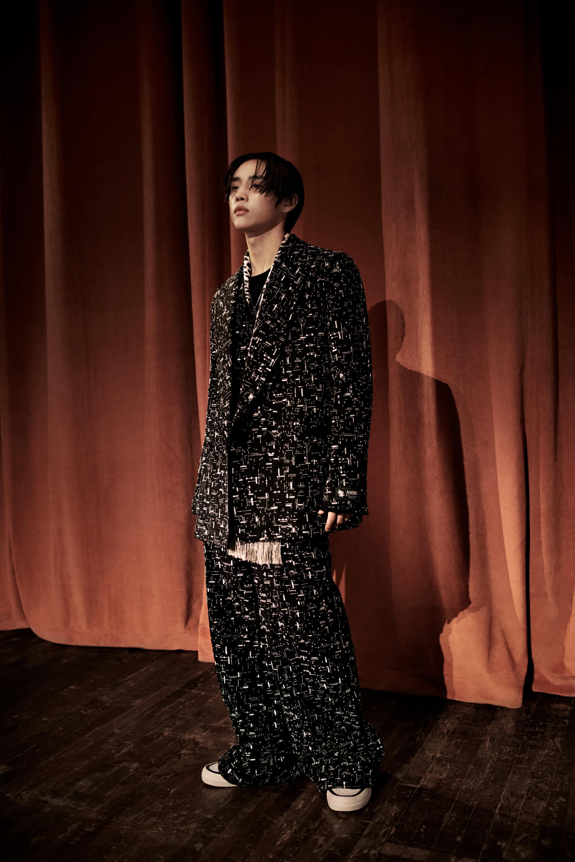 Man wearing a textured black patterned suit standing on a wooden stage against dark red velvet curtains.