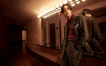 Man wearing a brown plaid suit and red shirt leaning against a mirror with Hollywood vanity lights in a hallway.