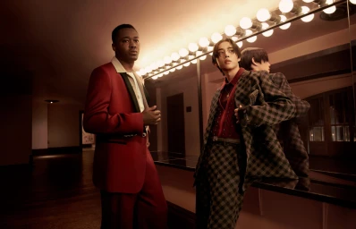 Two men in tailored suits posing in front of a mirror with warm vanity lighting for Amiri Fall 2024 ad campaign.