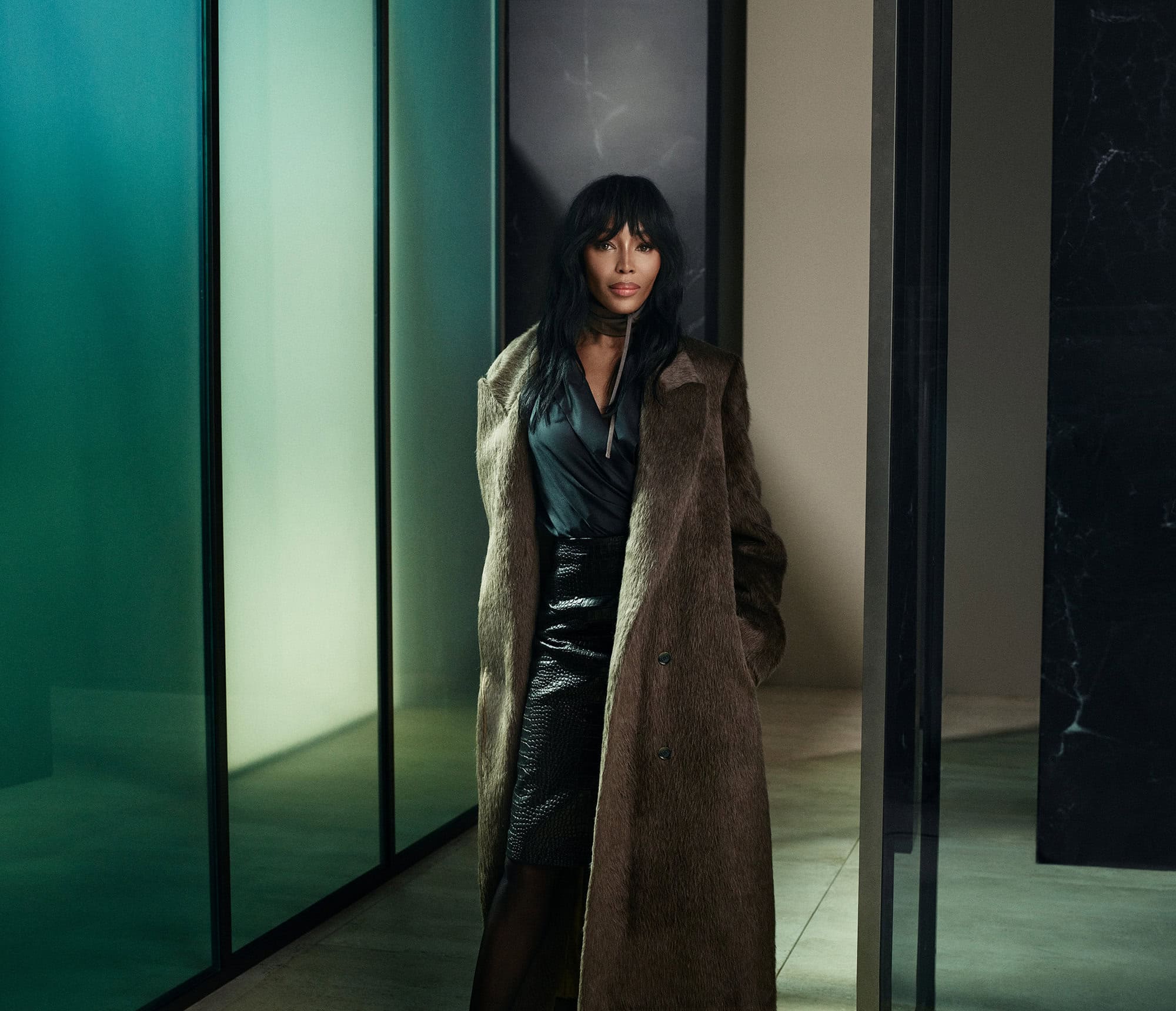 Naomi Campbell wearing a textured brown coat and dark skirt inside an architectural interior with glowing green panels