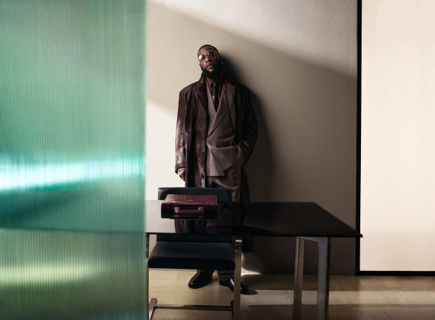 Burna Boy leaning against a wall in a brown suit and leather coat behind a dark desk with a green glass panel in the foreground.