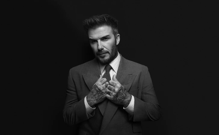 David Beckham in a dark tailored suit and tie against a plain black background for the BOSS Fall 2024 campaign.