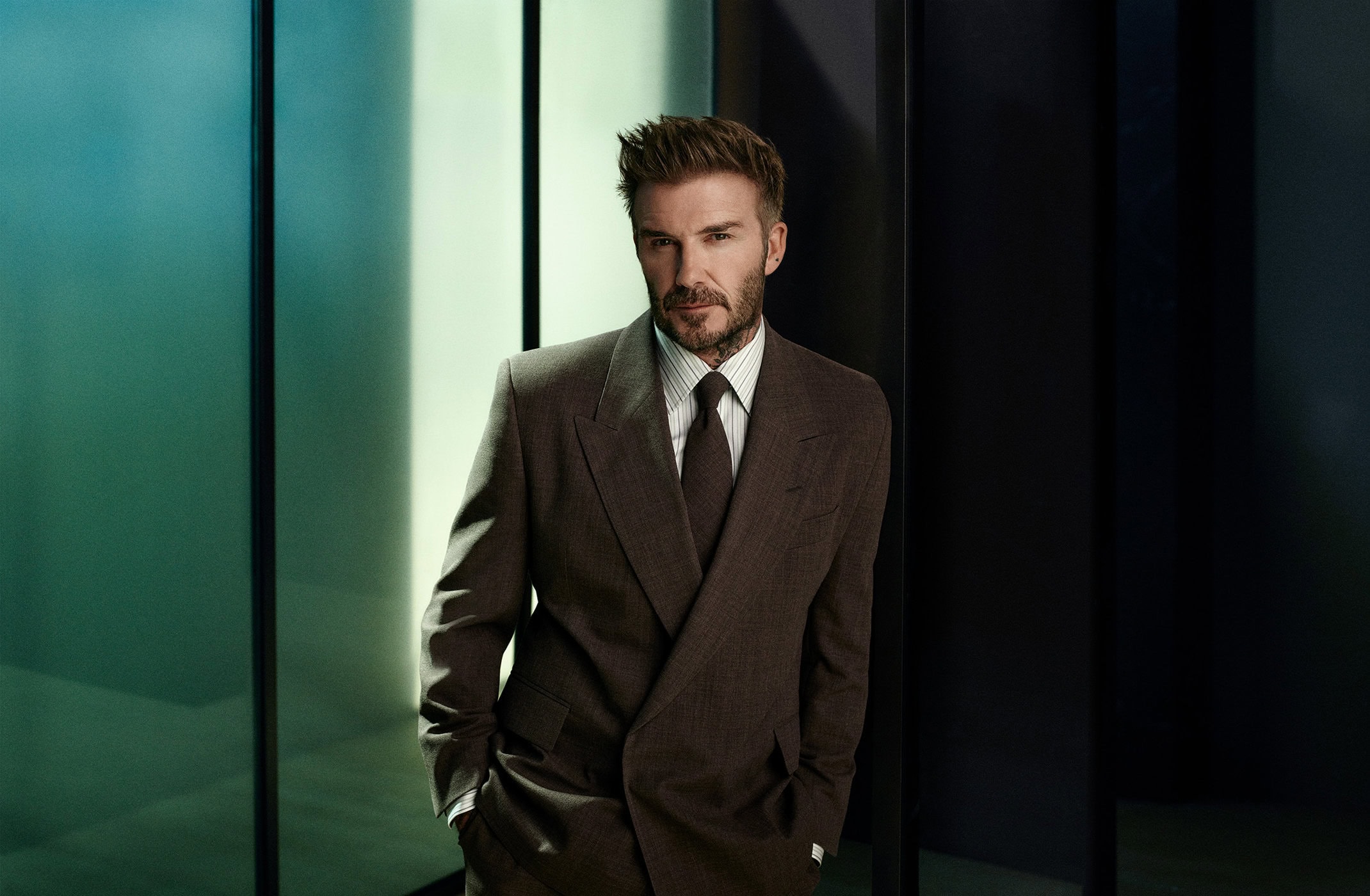 David Beckham wearing a brown double-breasted suit and tie against an illuminated glass wall background for BOSS Fall 2024 campaign.