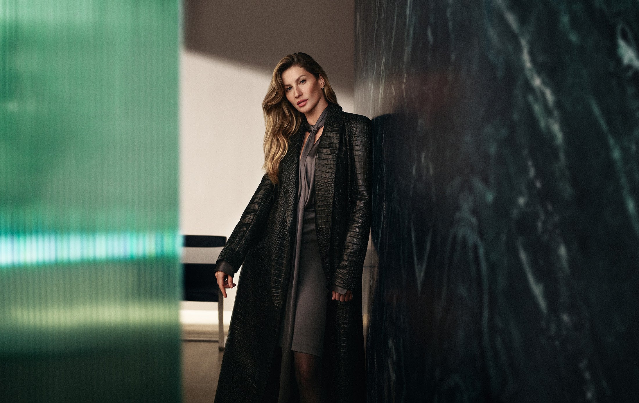 Gisele Bundchen wearing a long black embossed coat leaning against a dark marble wall for BOSS Fall 2024 campaign
