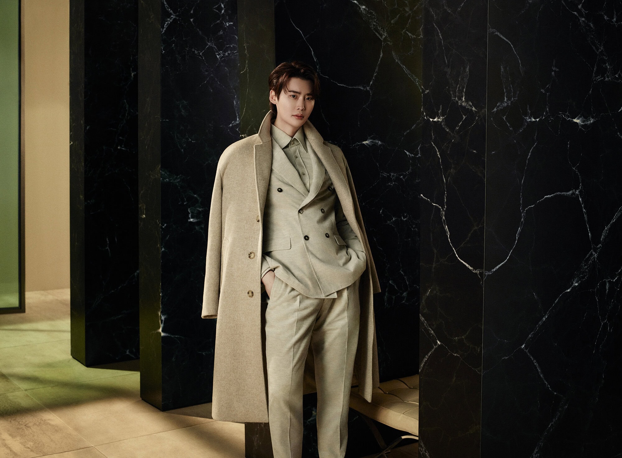 Lee Jong-suk wearing a camel coat and beige suit standing against a dark marble wall for the BOSS Fall 2024 campaign.