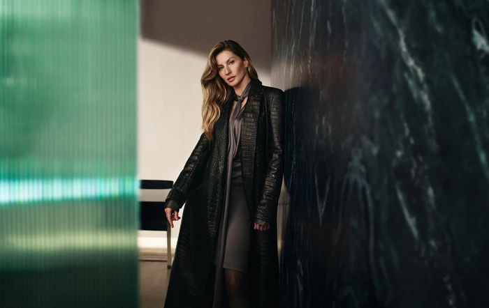 Gisele Bundchen wearing a long black embossed coat leaning against a dark marble wall for BOSS Fall 2024 campaign