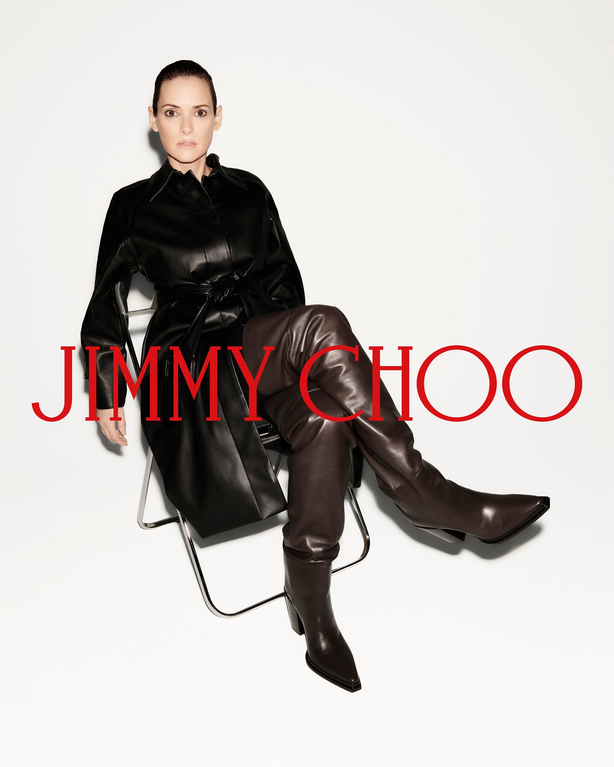 Winona Ryder seated on a wire frame chair wearing a black leather coat and brown leather boots against a white background