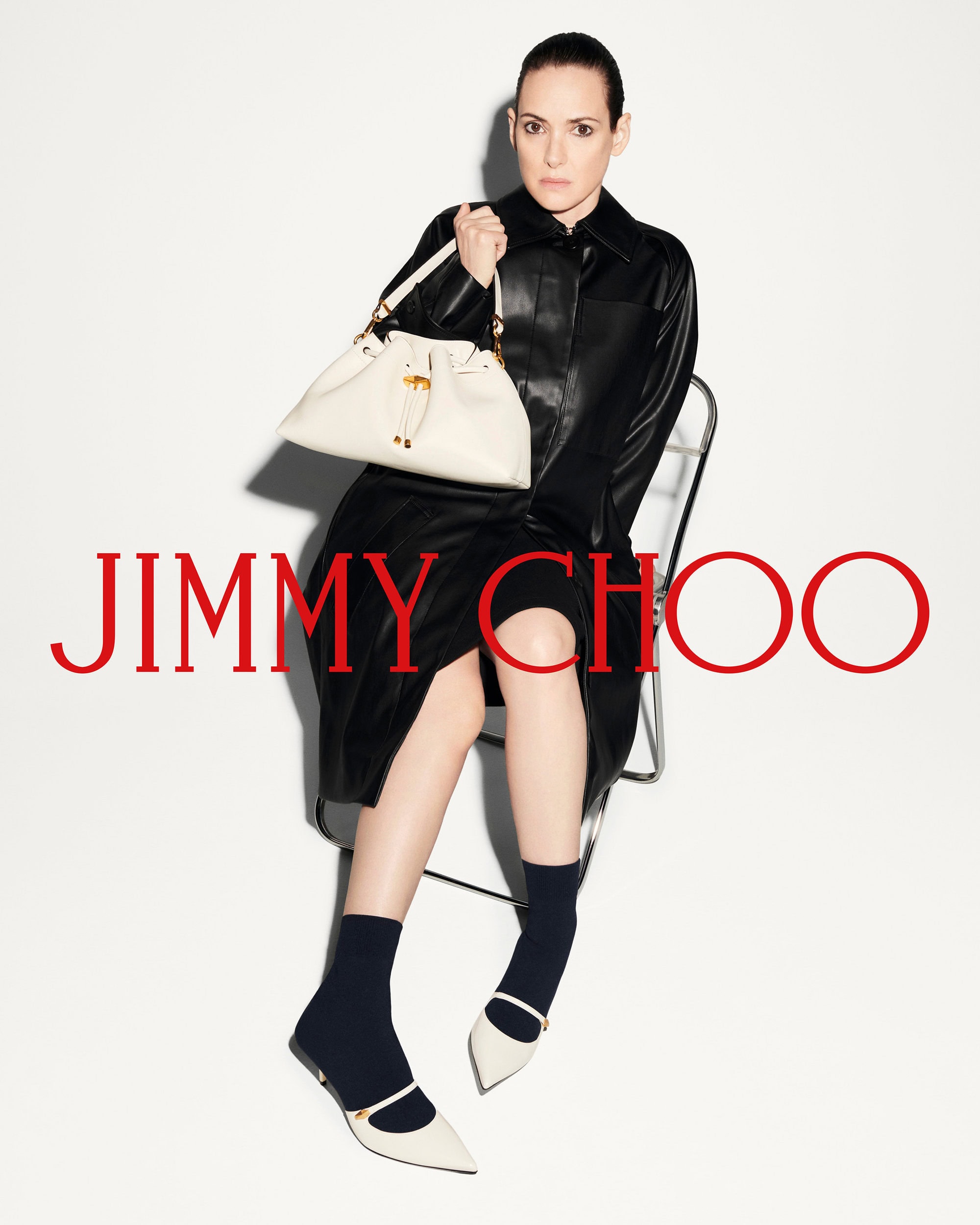 Winona Ryder seated on a chair wearing a black leather coat and holding a white handbag against a seamless white background
