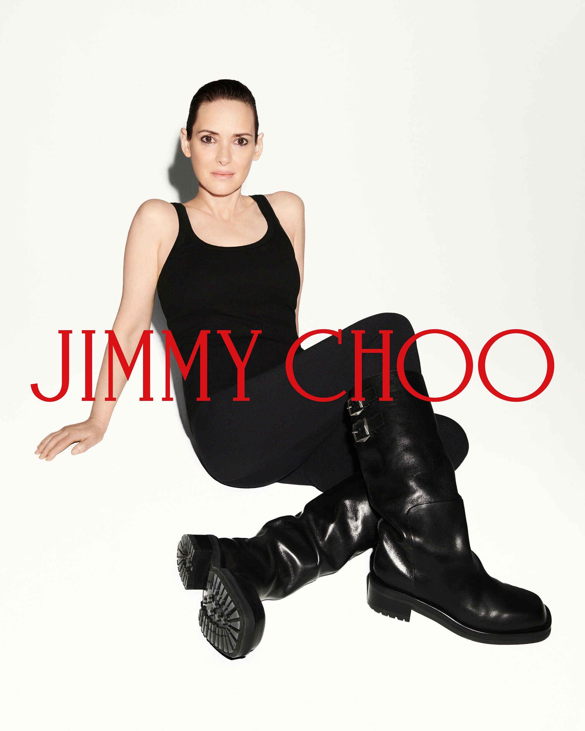 Winona Ryder seated against a white background wearing a black tank top and tall black leather boots for Jimmy Choo.