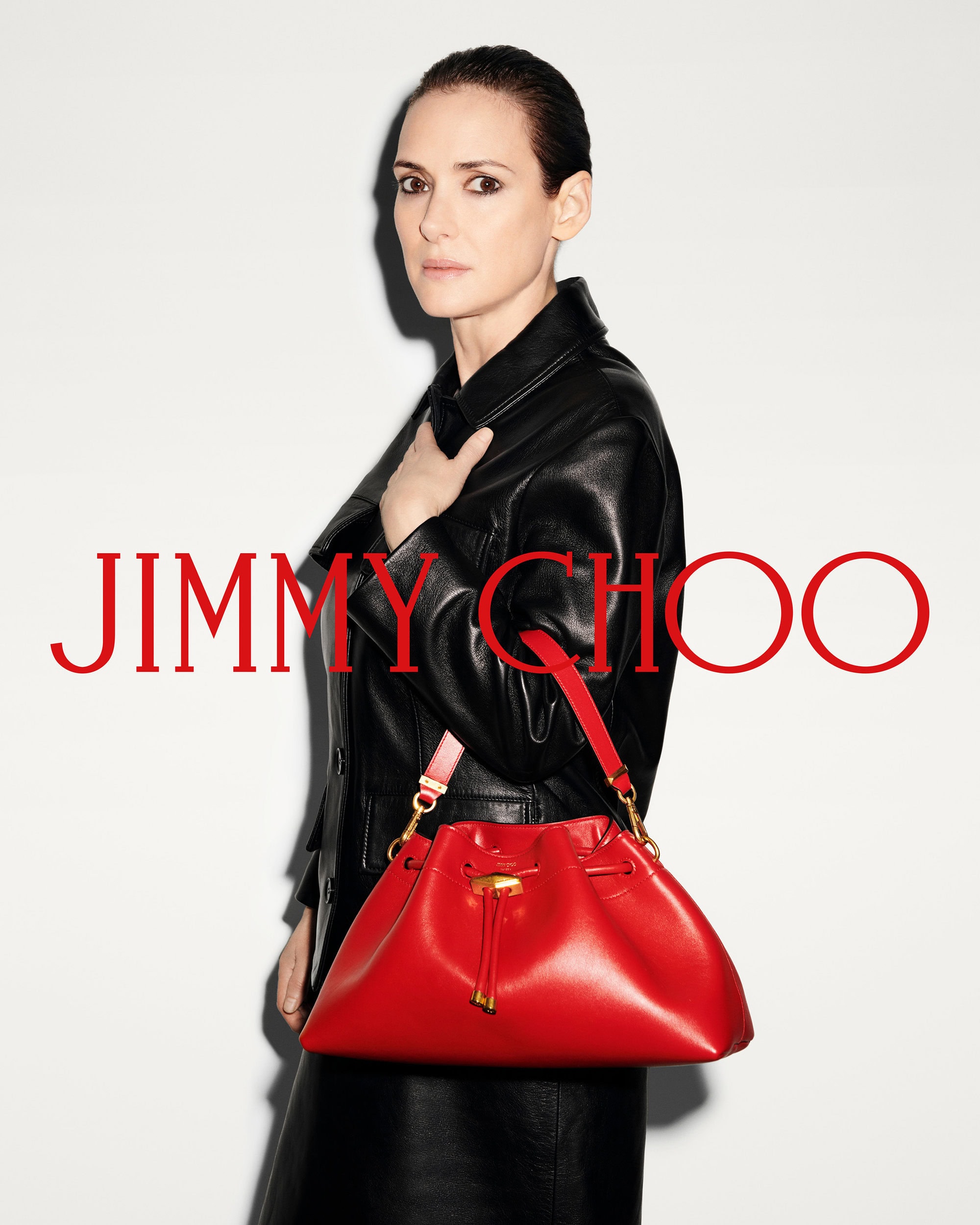 Winona Ryder wearing a black leather coat holding a bright red bucket bag against a plain white studio background.
