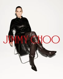 Winona Ryder seated on a wire frame chair wearing a black leather coat and brown leather boots against a white background