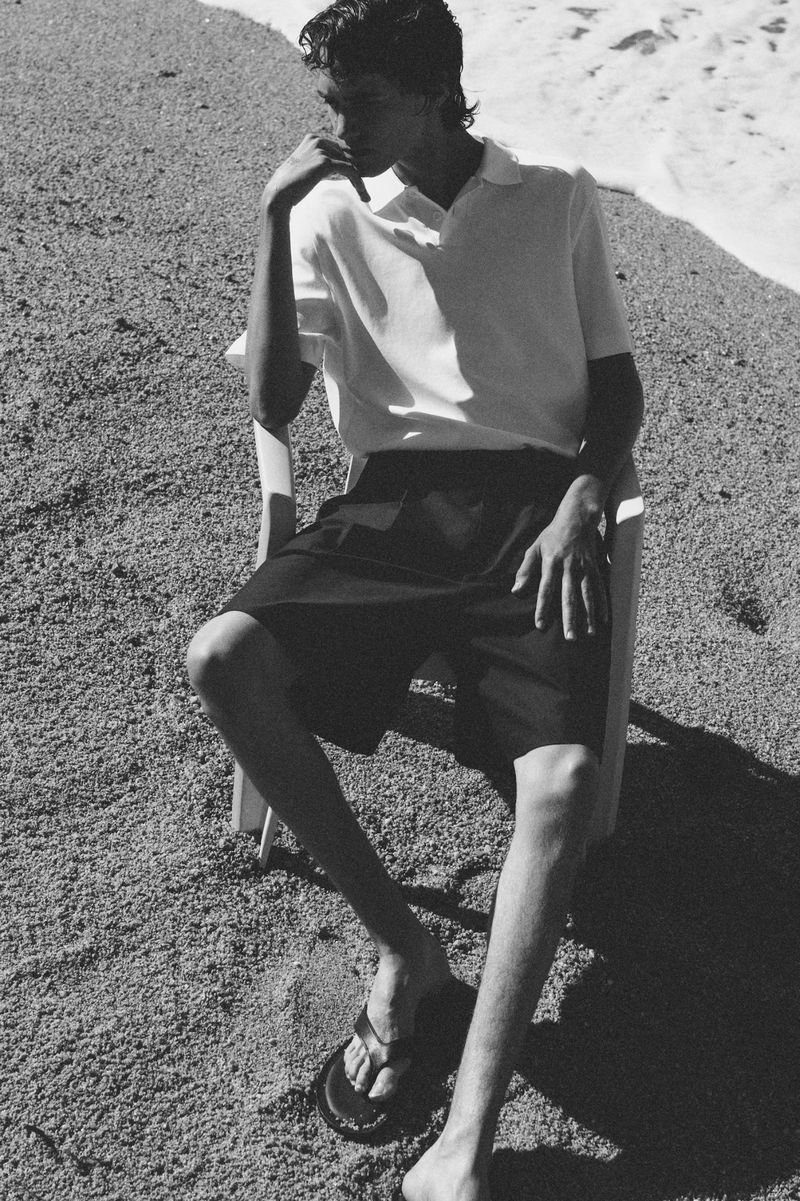 Monochrome portrait of a male model wearing a polo shirt and shorts, sitting on a chair at the beach near the ocean on a sunny day.