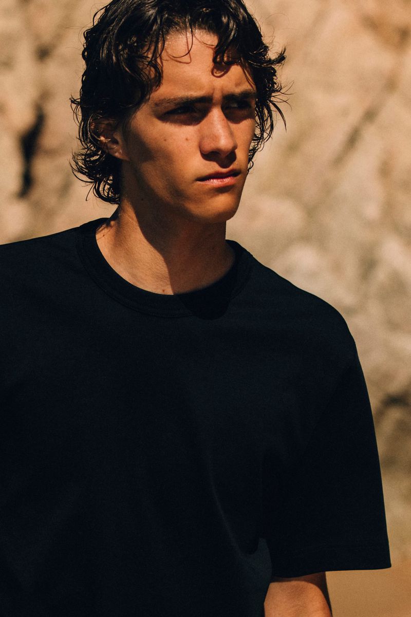 Portrait of a young adult male in a black t-shirt against a beige stone background, featuring natural lighting and a medium shot.