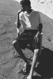 Model seated in a plastic chair on a sandy beach wearing a white polo shirt and dark shorts