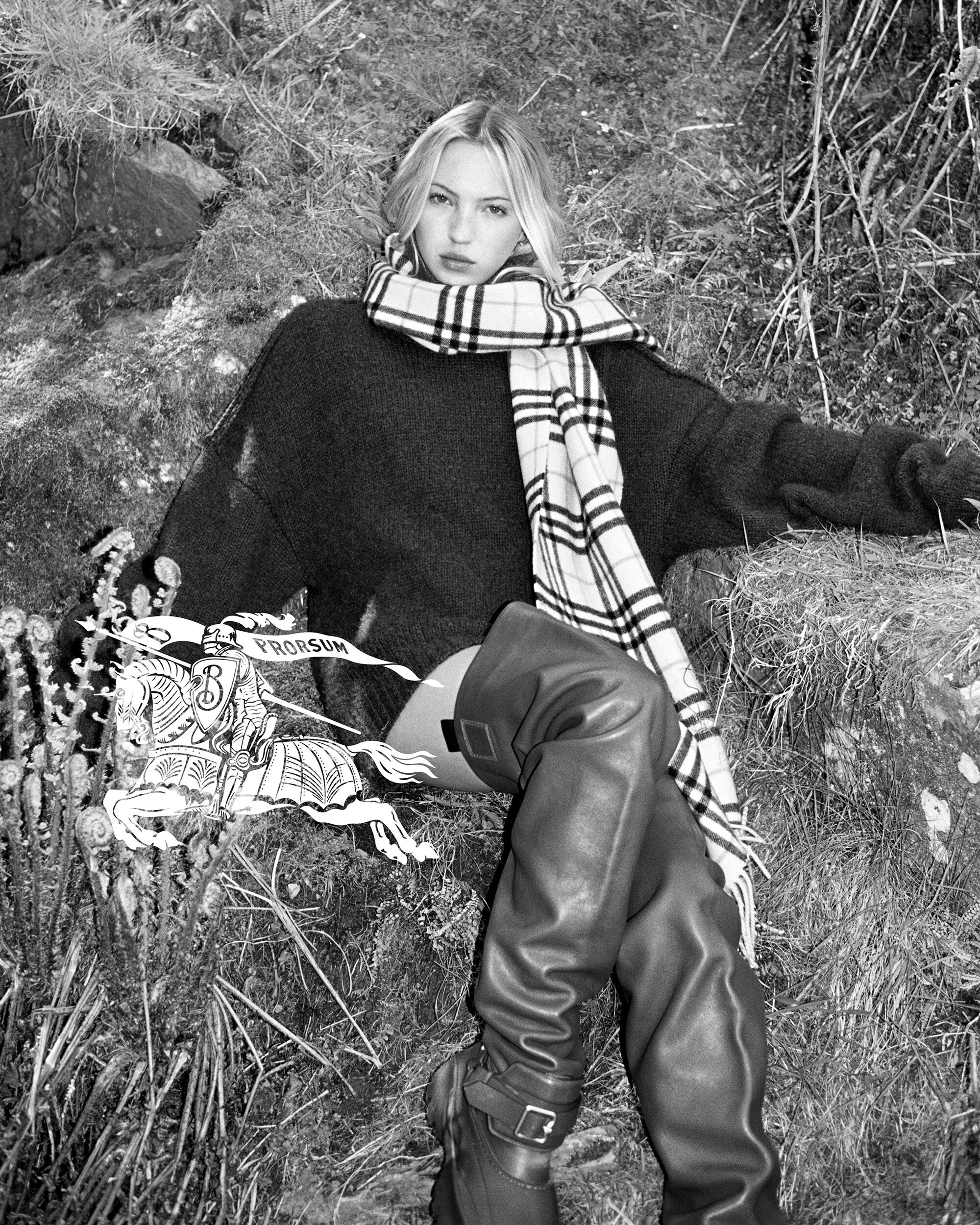 Model wearing a plaid scarf and dark sweater seated outdoors among rocky terrain and ferns in a black and white editorial photograph.