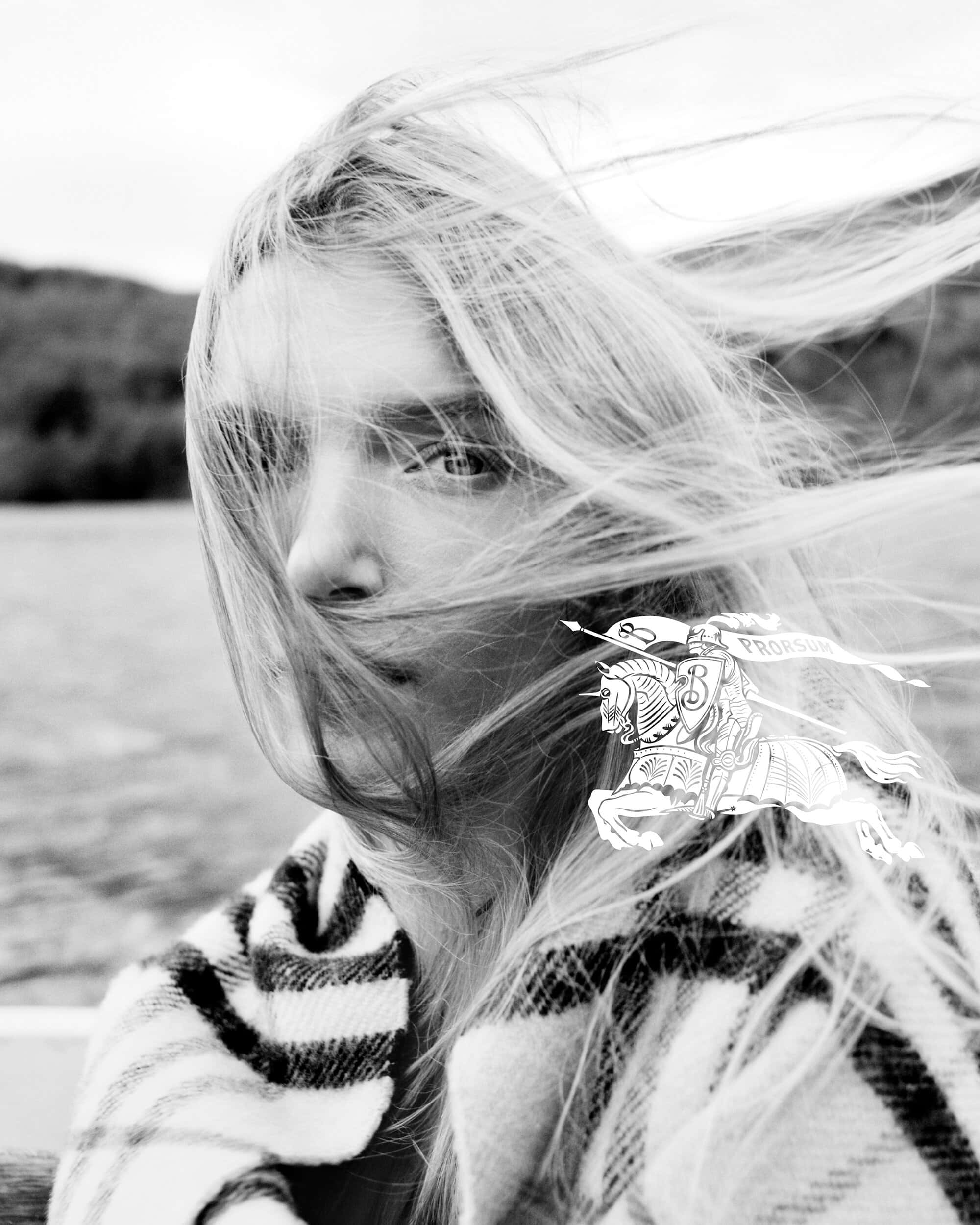 Close-up portrait of a model with blonde hair blowing across her face wearing a plaid scarf outdoors.