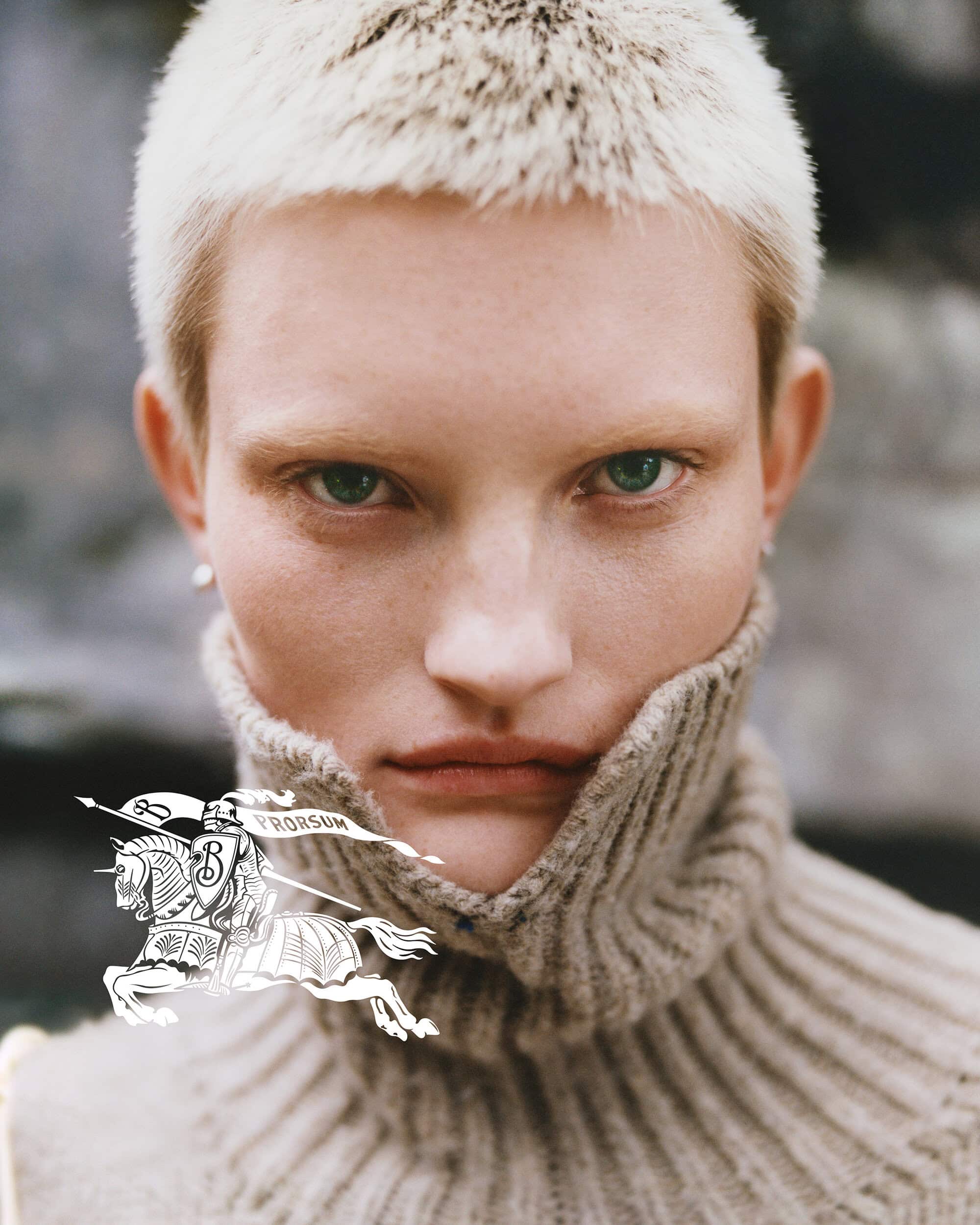 Close-up portrait of a model with short blonde hair and green eyes wearing a heavy beige turtleneck sweater against an outdoor background.