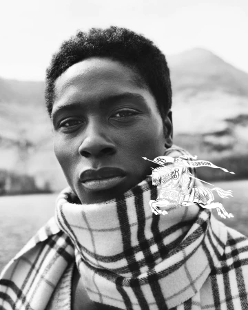 Portrait of a Black man wearing a plaid scarf and jacket outdoors against a mountain background in a monochrome photograph