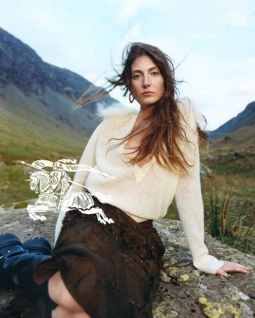 Model in cream knit sweater and dark skirt seated outdoors against a mountainous landscape for Burberry