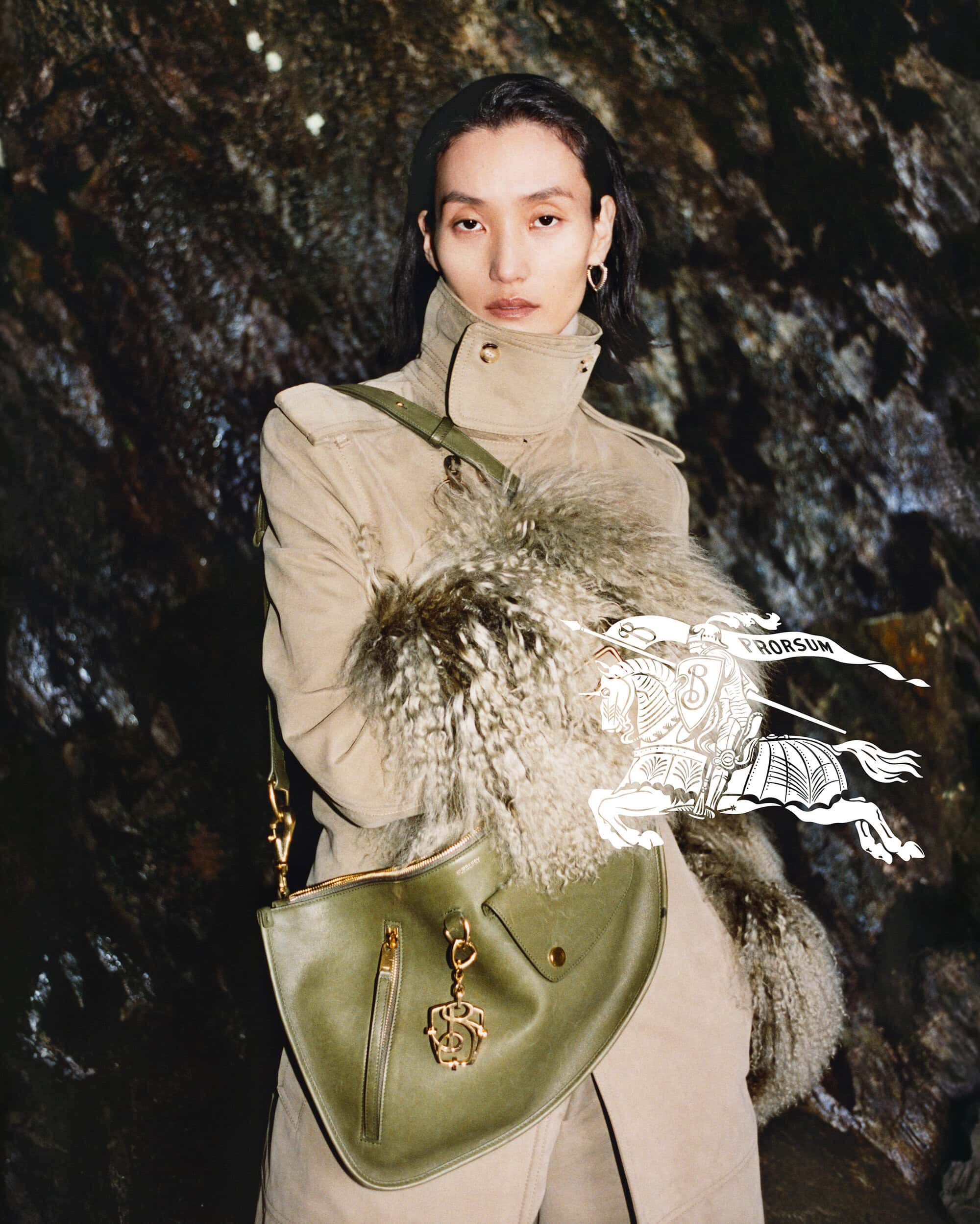 Model wearing a beige coat and holding an olive green handbag against a rocky background