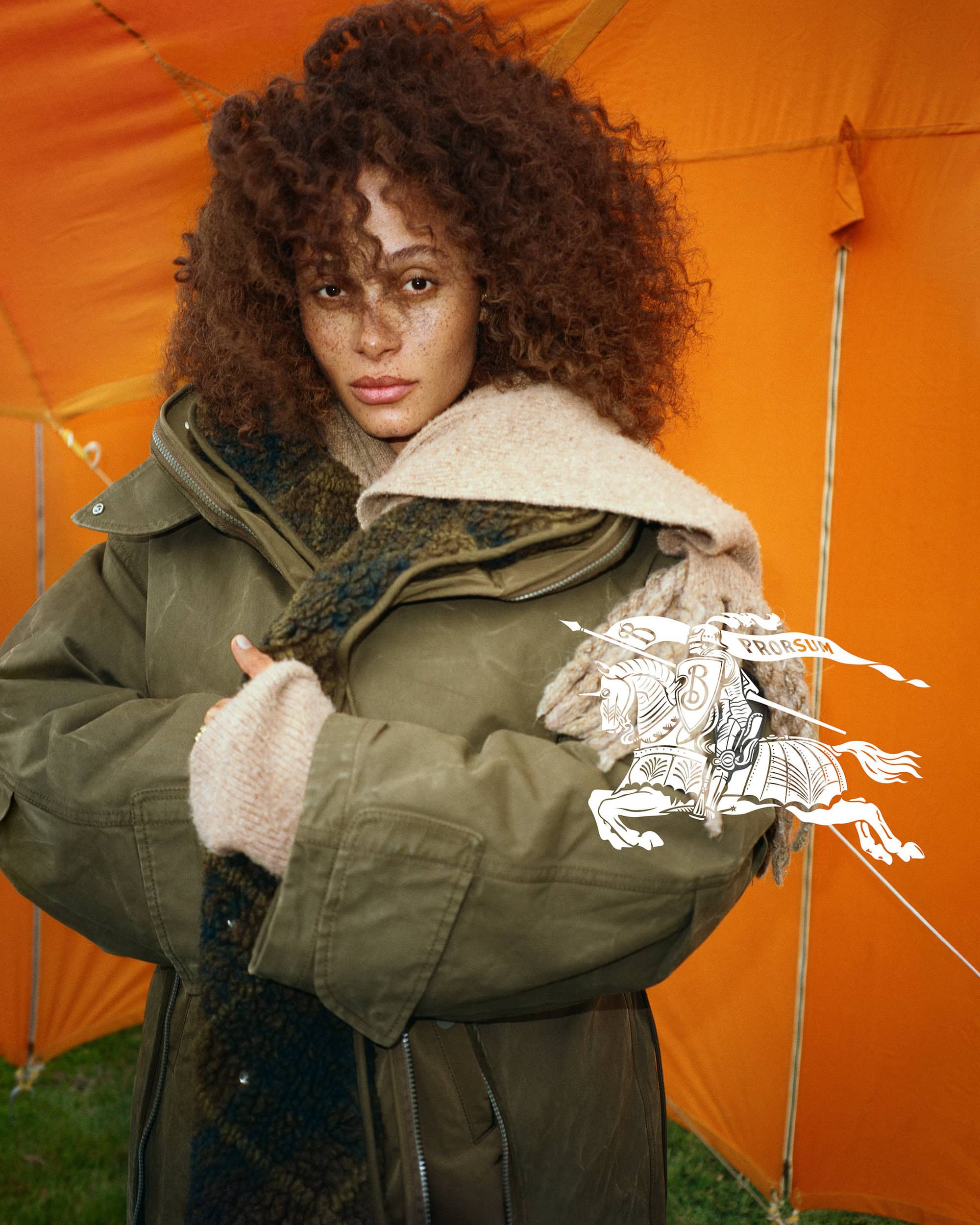 Model in olive green coat and textured scarf standing against an orange tent backdrop for Burberry Winter 2024 campaign