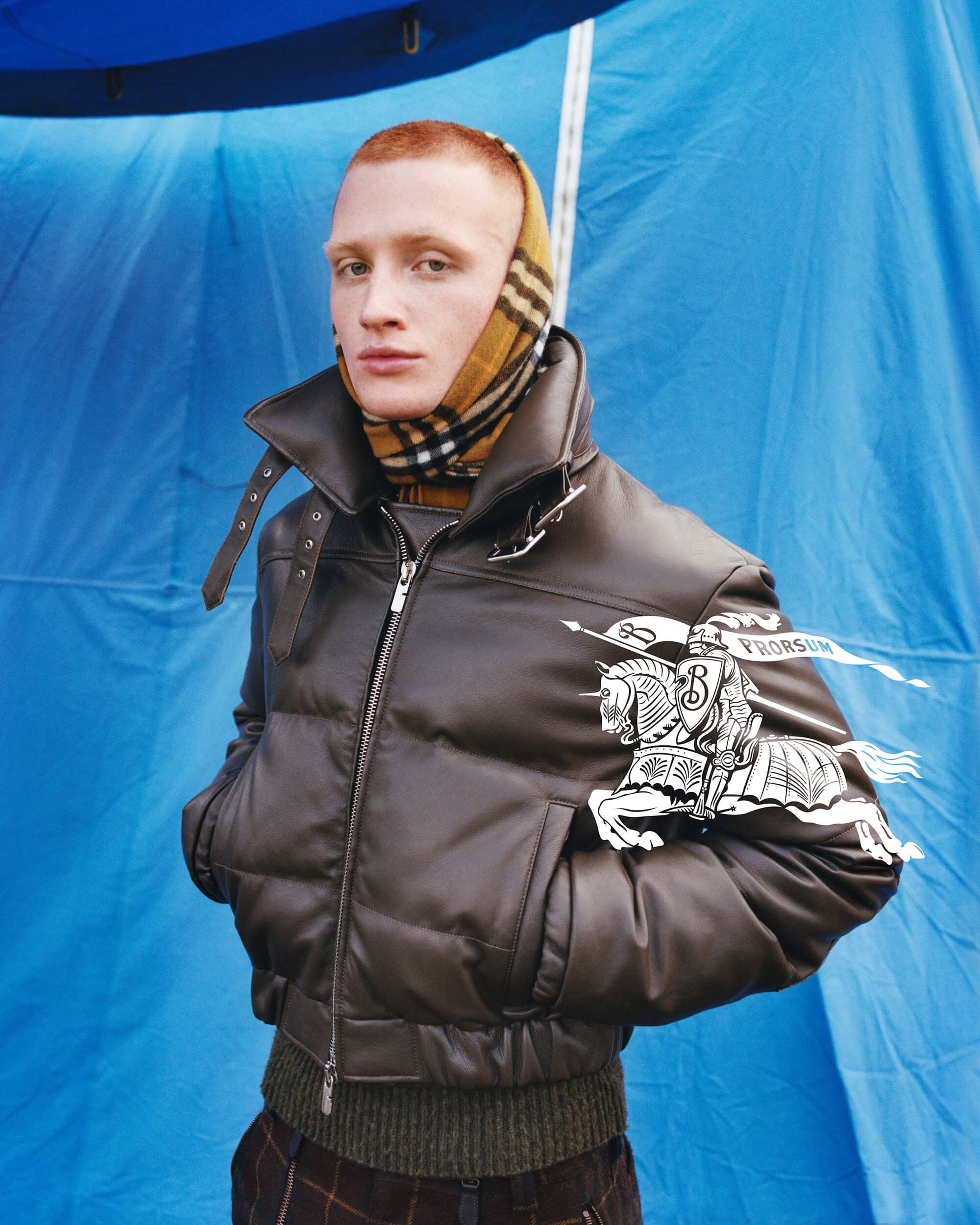 Man wearing a brown leather puffer jacket and plaid scarf against a blue tarp backdrop for Burberry Winter 2024 campaign